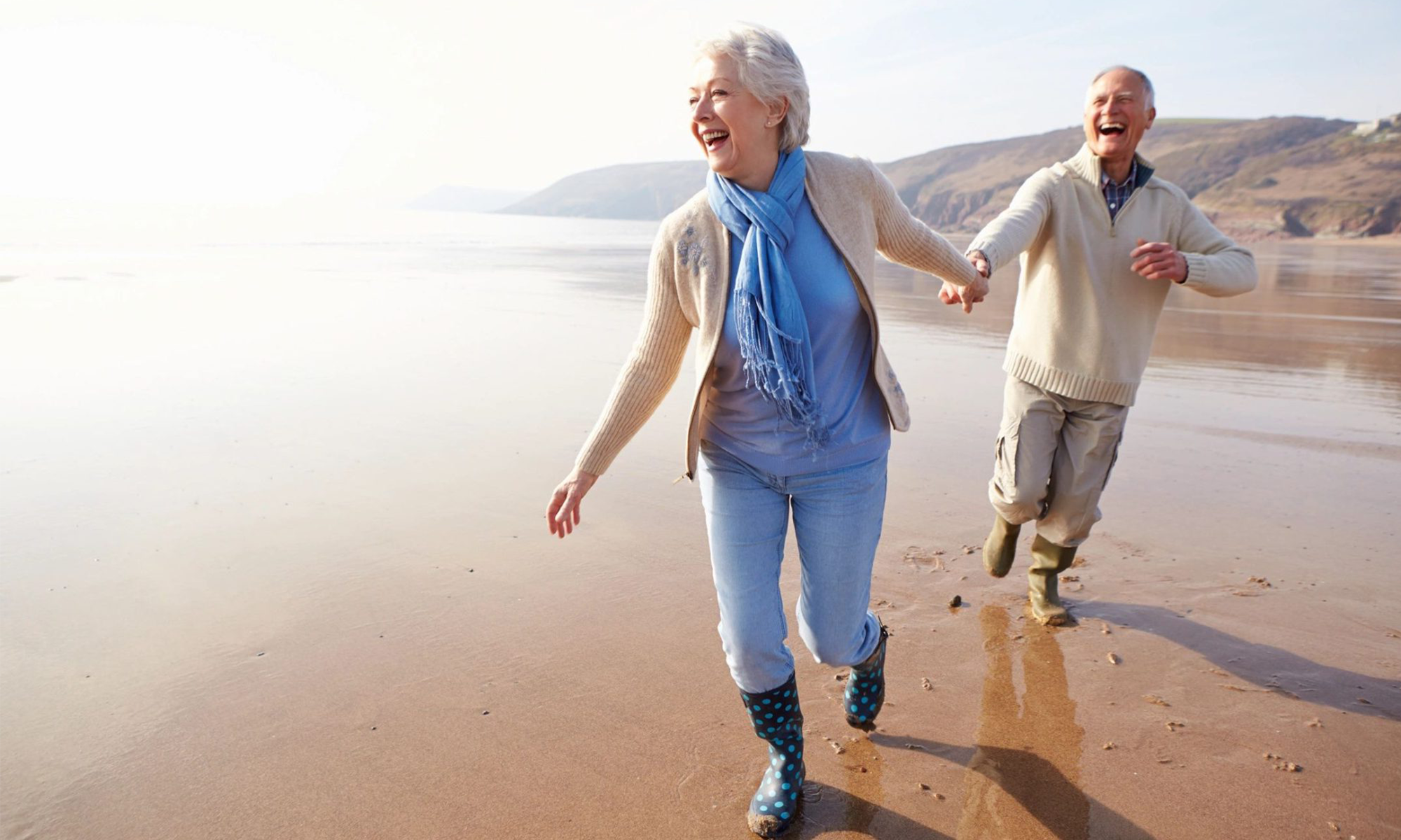 An elderly woman pulling her husband along behind her as they run along a beach, laughing