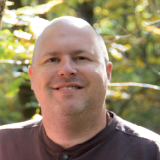 Head shot of Steve Fuller, smiling, standing in front of green trees